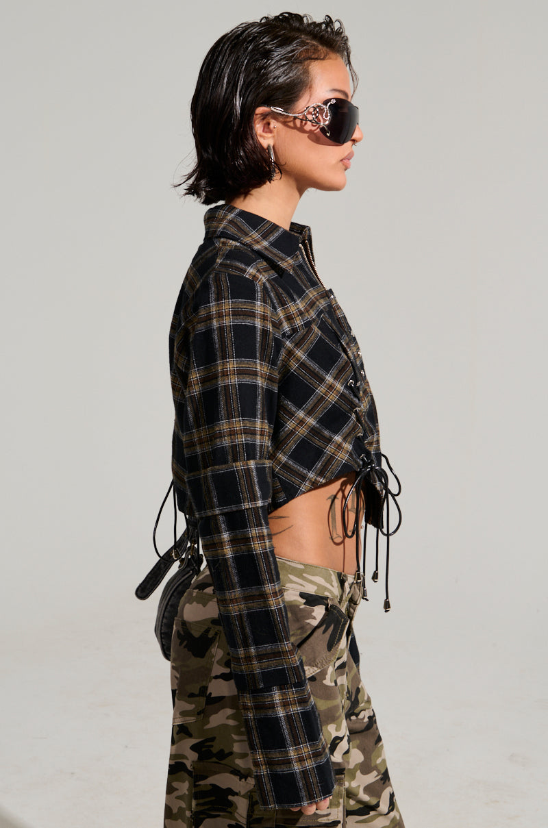 A person with short dark hair is wearing oversized sunglasses, an UNSPEAKABLE BUTTON DOWN PLAID SHIRT featuring a cropped silhouette in black and gray, along with camouflage pants. They have a small black bag and are standing against a plain background, facing to the side.