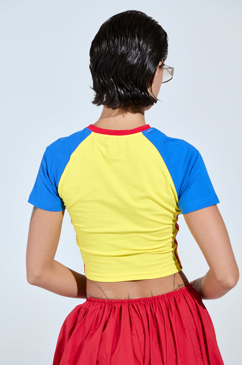 A person with short dark hair faces away, wearing the COLOMBIA BABY TEE featuring a yellow and blue color block design with red trim and "COLOMBIA" embroidered on the back. They sport a red skirt, revealing back tattoos against a plain, light-colored background.