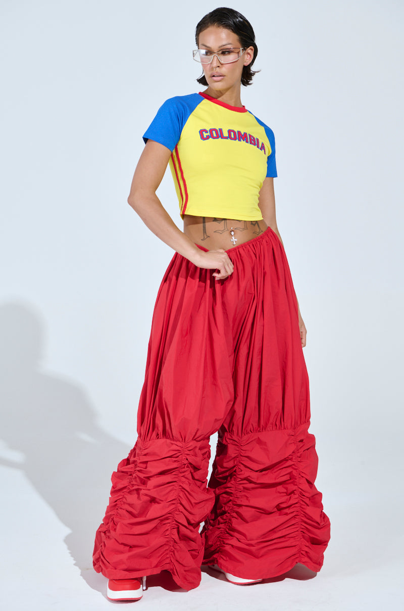 Wearing a COLOMBIA BABY TEE with bold red voluminous ruched pants and clear glasses, a person strikes a pose against a plain white background.