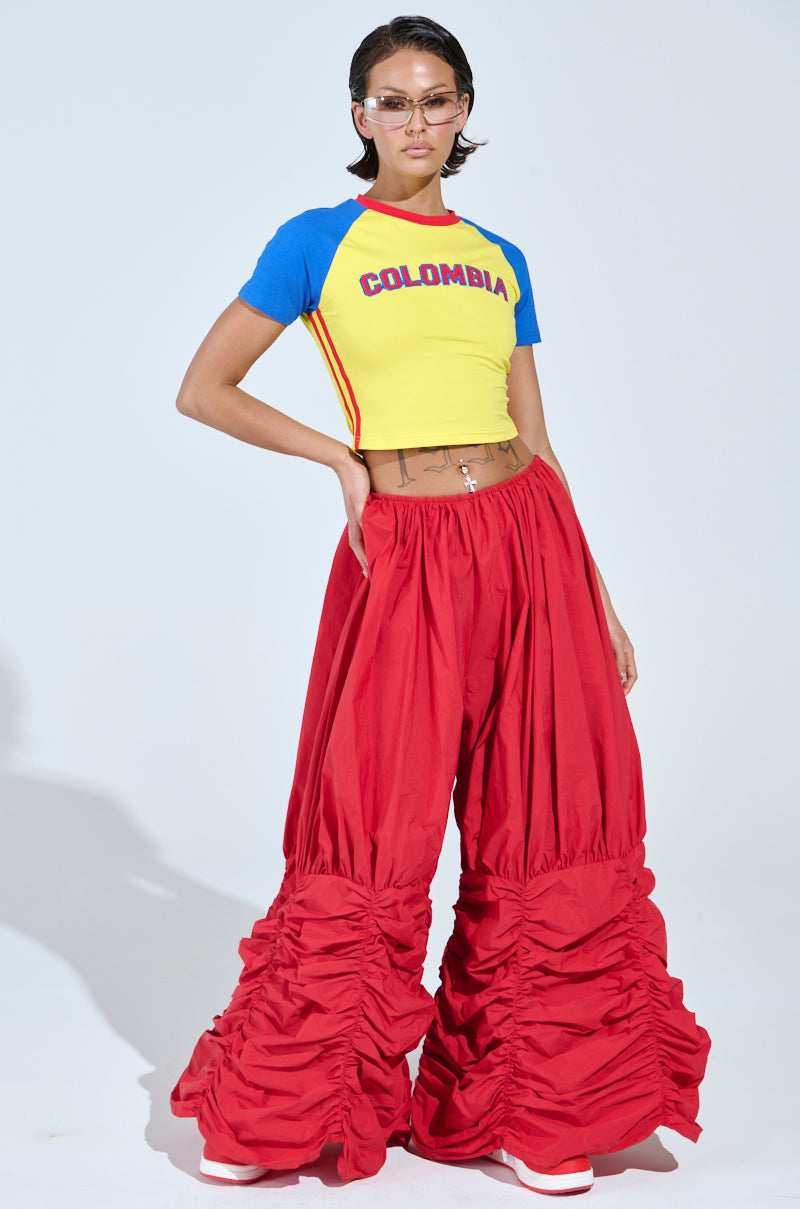 A person confidently models the COLOMBIA BABY TEE, an embroidered yellow crop top with "COLOMBIA." Styled with oversized red pants featuring gathered details, they exude flair with short hair, glasses, and stomach tattoos.