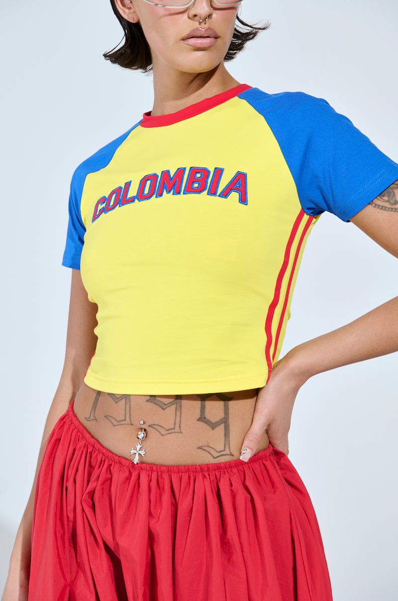 Wearing a bold color block COLOMBIA BABY TEE with red stripes and "Colombia" embroidered, paired with a striking red skirt, the person completes their stylish look with glasses and tattoos on the midriff.