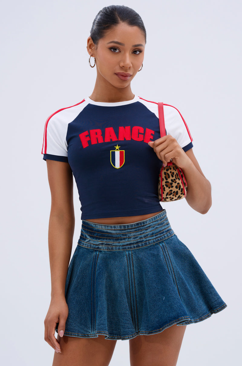 A woman poses against a plain backdrop wearing the FRANCE BABY TEE, a navy crop top with red lettering and a crest, paired with a short denim skirt and holding a small leopard-print purse.