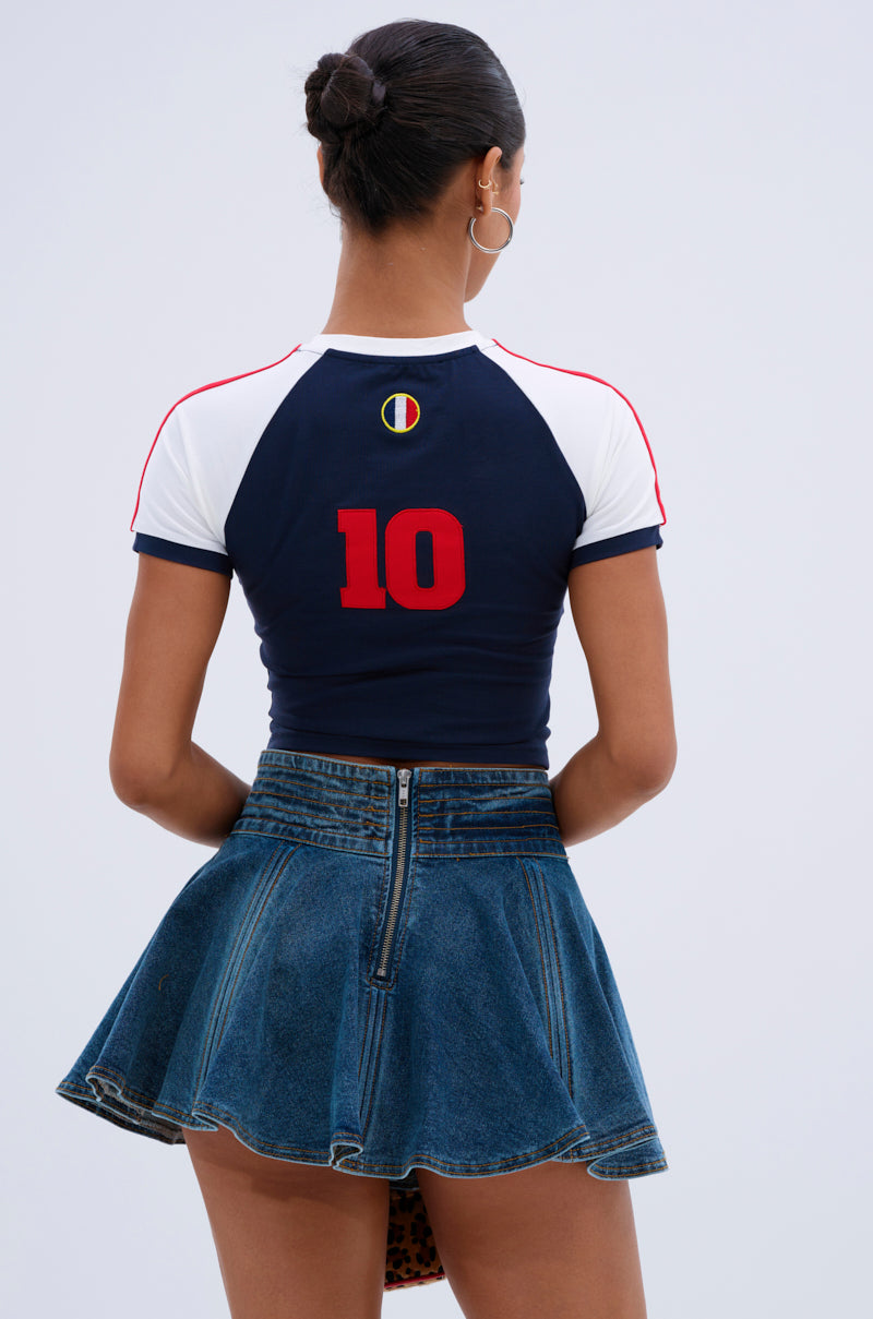 A woman faces away, wearing the FRANCE BABY TEE featuring a cropped silhouette, navy and white color block with red piping, a red number 10 on the back, and a high-waisted blue denim skirt with a visible back zipper.