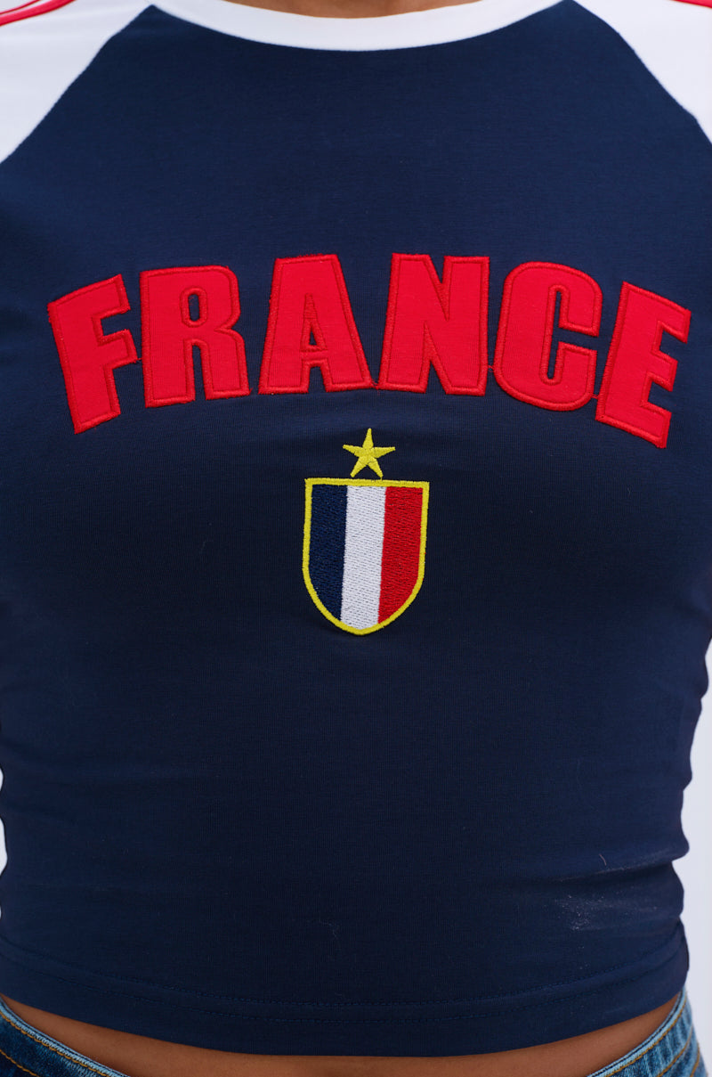 A close-up of the FRANCE BABY TEE, a navy blue cotton color block shirt with "FRANCE" in bold red letters above a blue, white, and red shield logo topped by a gold star.