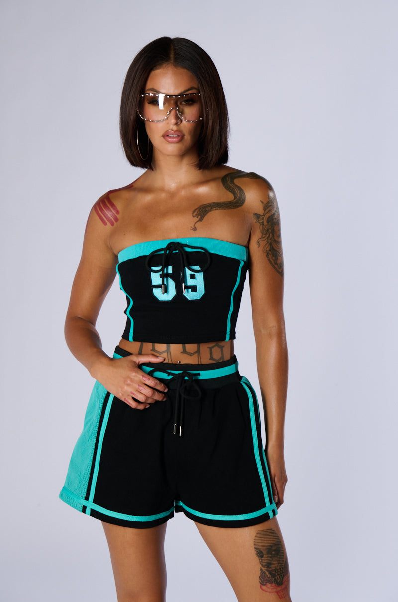 A woman with sunglasses and tattoos poses confidently in a matching black and turquoise BALLIN ALL NIGHT BANDEAU TOP and shorts set. The outfit, crafted from a cotton-blend fabrication, complements her short bob hairstyle as she stands against a plain background. 1