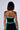 A person with short, straight hair is seen from behind, wearing a strapless BALLIN ALL NIGHT BANDEAU TOP with matching bottoms in black and turquoise. They have tattoos on their upper back and left arm, along with three parallel red marks on their right shoulder. The background is plain gray.