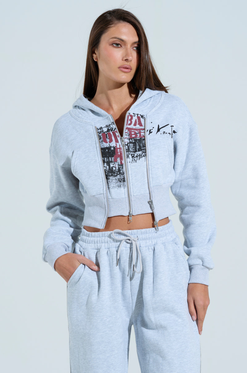 A person in a light grey tracksuit features the WILD AND FREE SWEATSHIRT with graffiti-inspired graphics and drawstring pants, stands with one hand in a pocket against a plain backdrop, exuding confidence and embodying the vibrant style of the AKIRA Label.