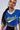 A person with long, braided dark hair is wearing a blue IN THE MOOD TEE featuring "Mood" in yellow script across the front. They are also sporting large hoop earrings, a chunky chain necklace, and black and white striped pants—an effortlessly sporty chic ensemble.