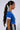 A woman with long, sleek, dark hair styled in a high ponytail is seen in profile. She is wearing the blue IN THE MOOD TEE with white trim and a pair of black athletic pants featuring white stripes. She accessorizes with large hoop earrings and a chunky chain necklace.