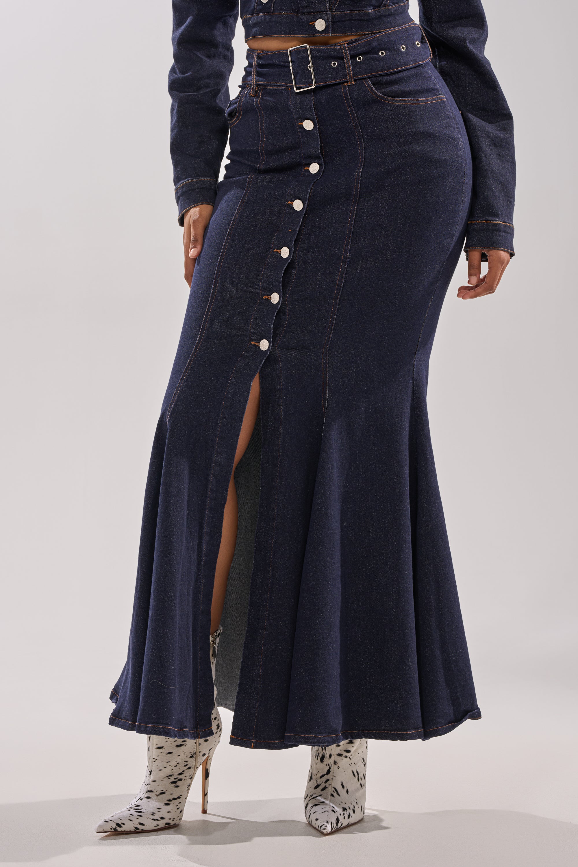Someone wears the YOU GOT ME SPINNING DENIM MAXI SKIRT, featuring a high-rise waist, front slit, button details, and matching belt, styled with a denim jacket and white pointed-toe boots with a black speckled pattern. 2