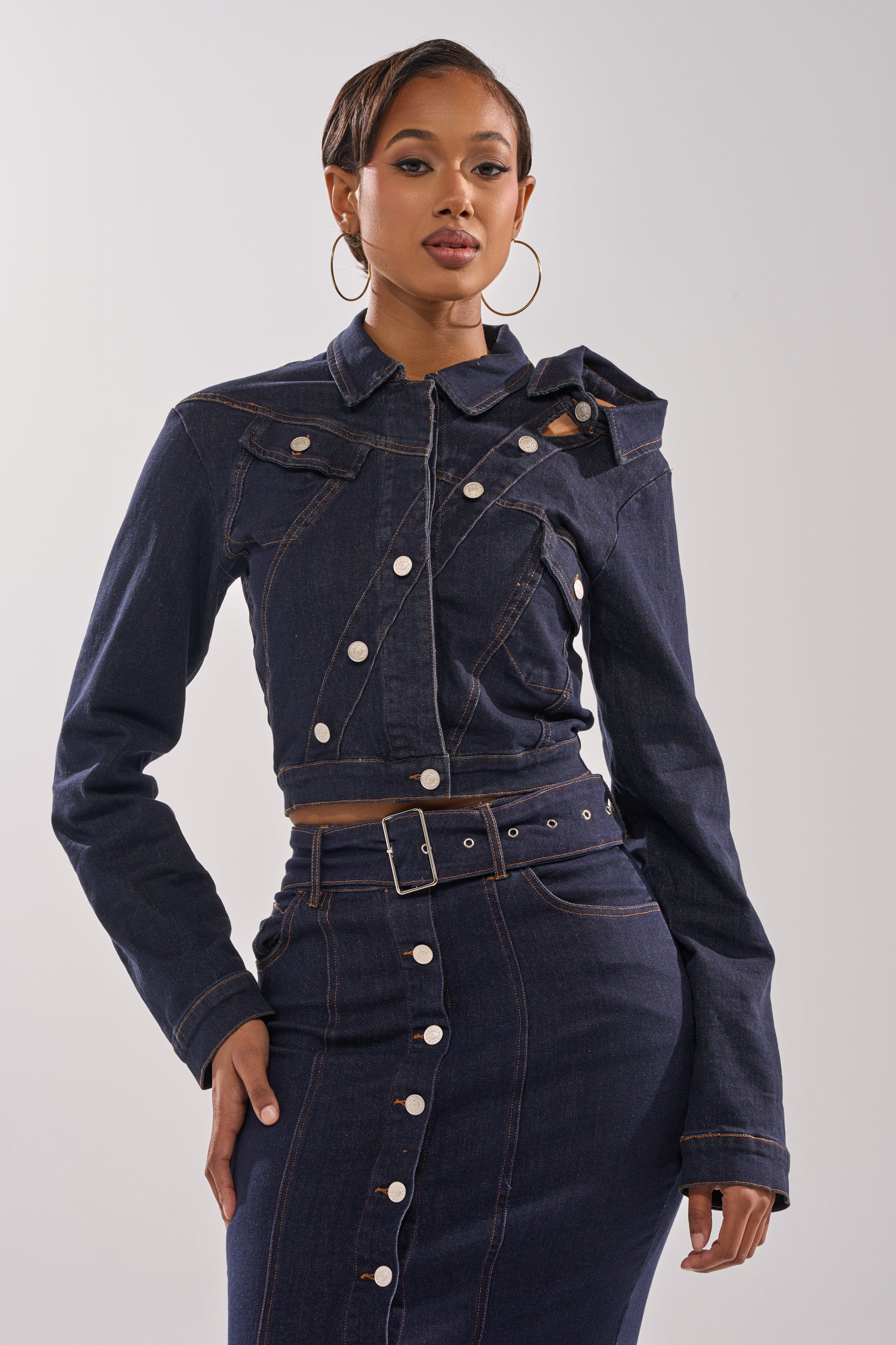 A woman models the YOU GOT ME SPINNING DENIM BUTTON DOWN, a cropped indigo jacket with asymmetrical buttons, paired with a matching belted high-waisted skirt, posing confidently against a plain backdrop.