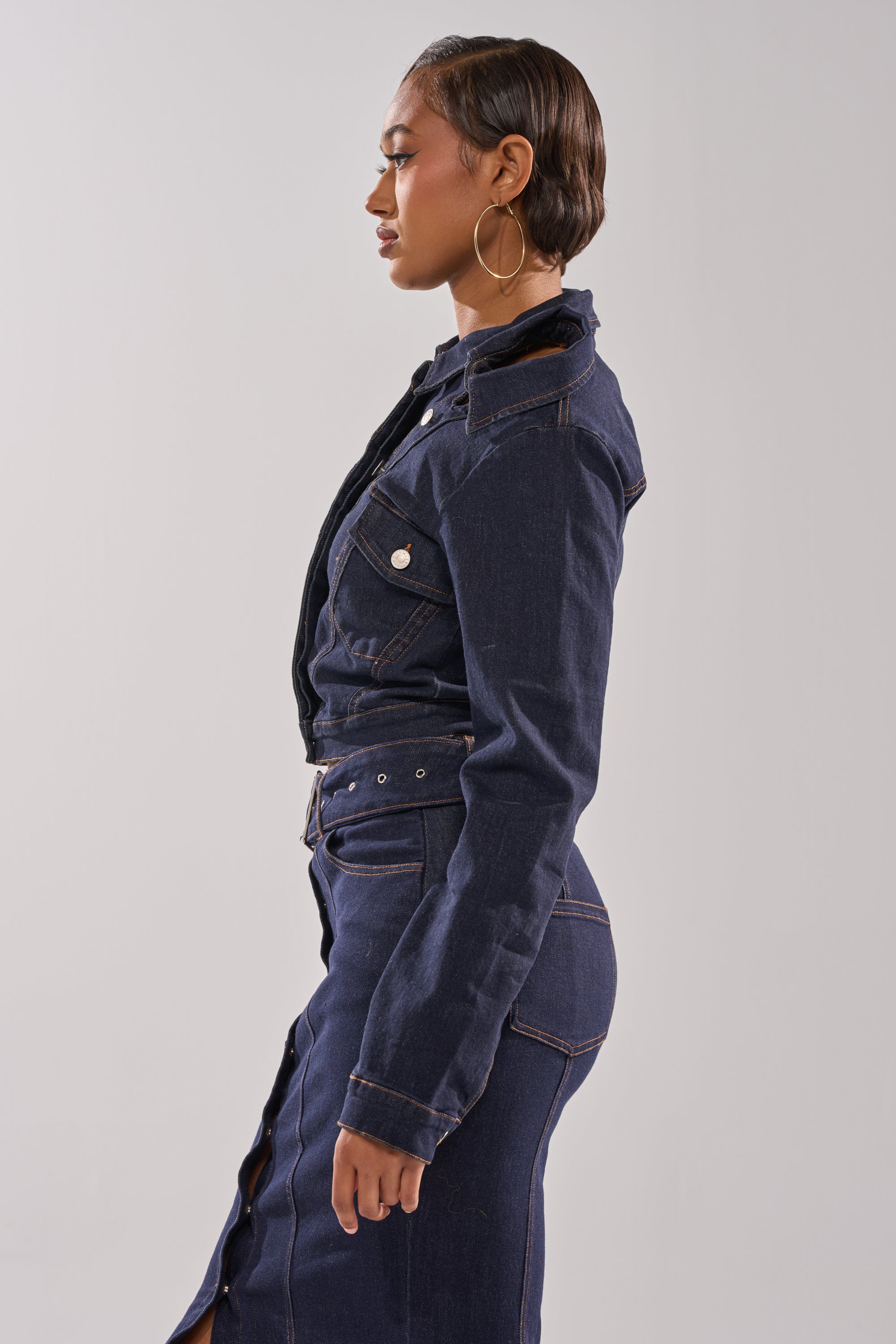A woman with short, slicked-back hair and large hoop earrings poses in profile wearing the YOU GOT ME SPINNING DENIM BUTTON DOWN, styled as a cropped jacket with an indigo wash and paired with a matching high-waisted skirt. The background is plain and light.