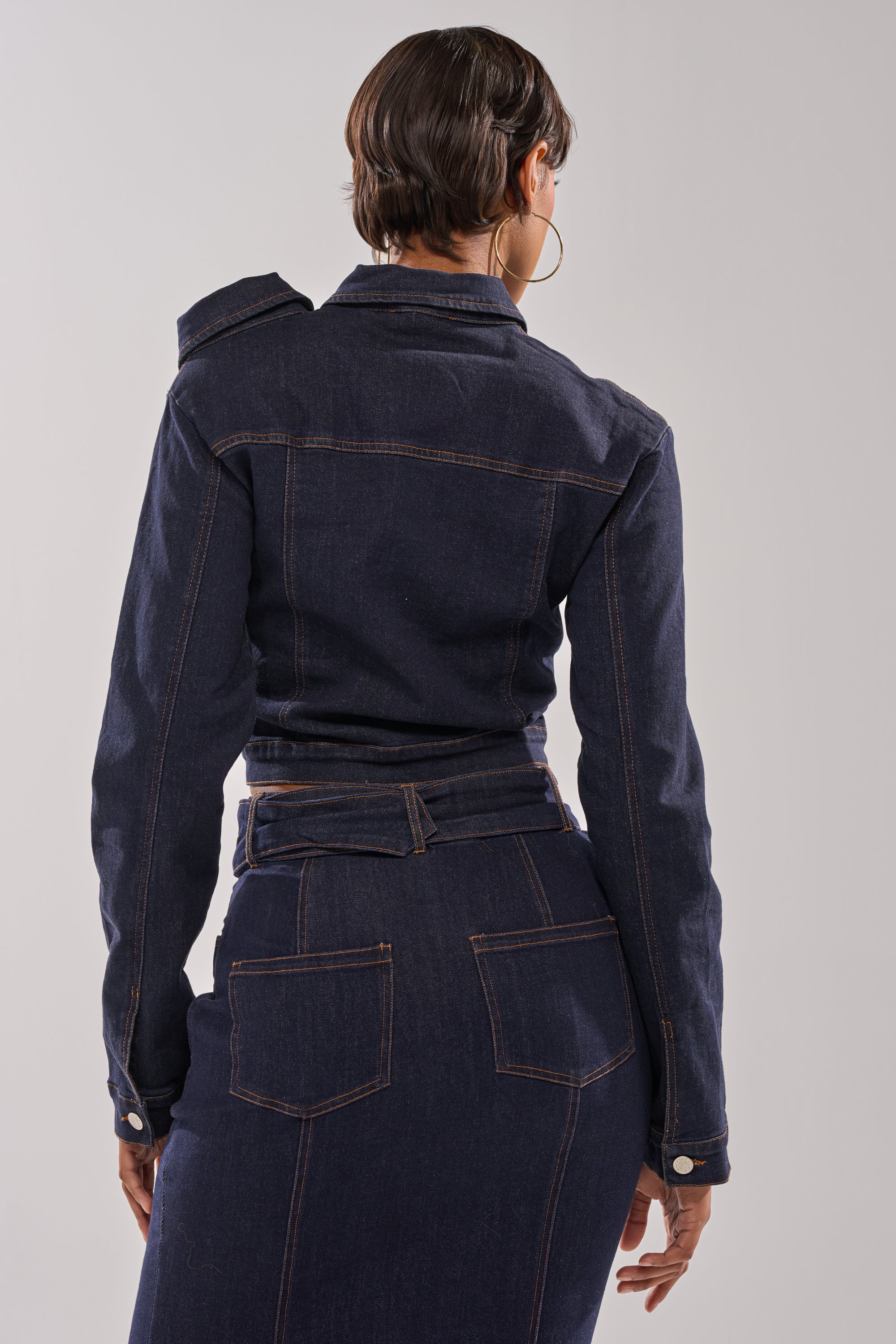 A person with short hair and large hoop earrings, wearing the YOU GOT ME SPINNING DENIM BUTTON DOWN as a fitted, cropped jacket with a matching high-waisted denim skirt, stands with their back to the camera against a plain background.