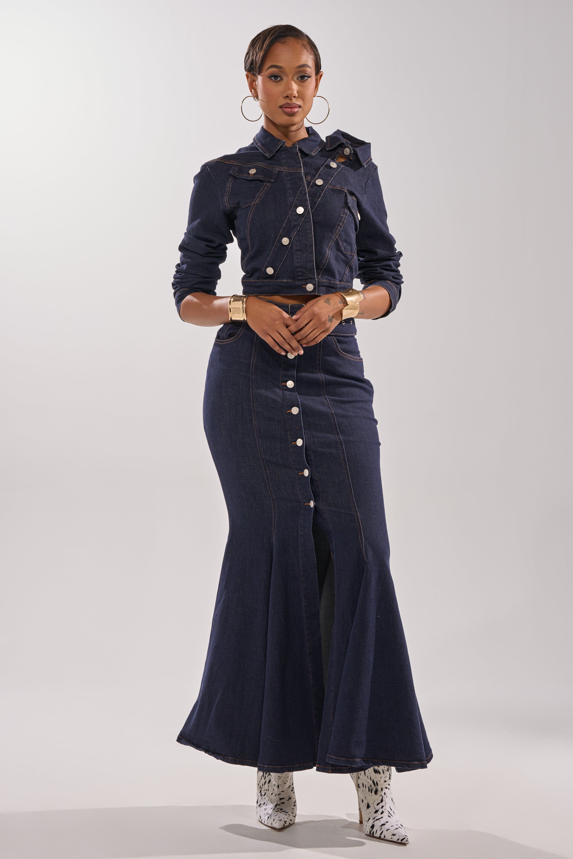 A woman stands confidently in the YOU GOT ME SPINNING DENIM BUTTON DOWN, an indigo wash fitted dress, styled with hoop earrings, gold bracelets, and white patterned ankle boots against a plain background.