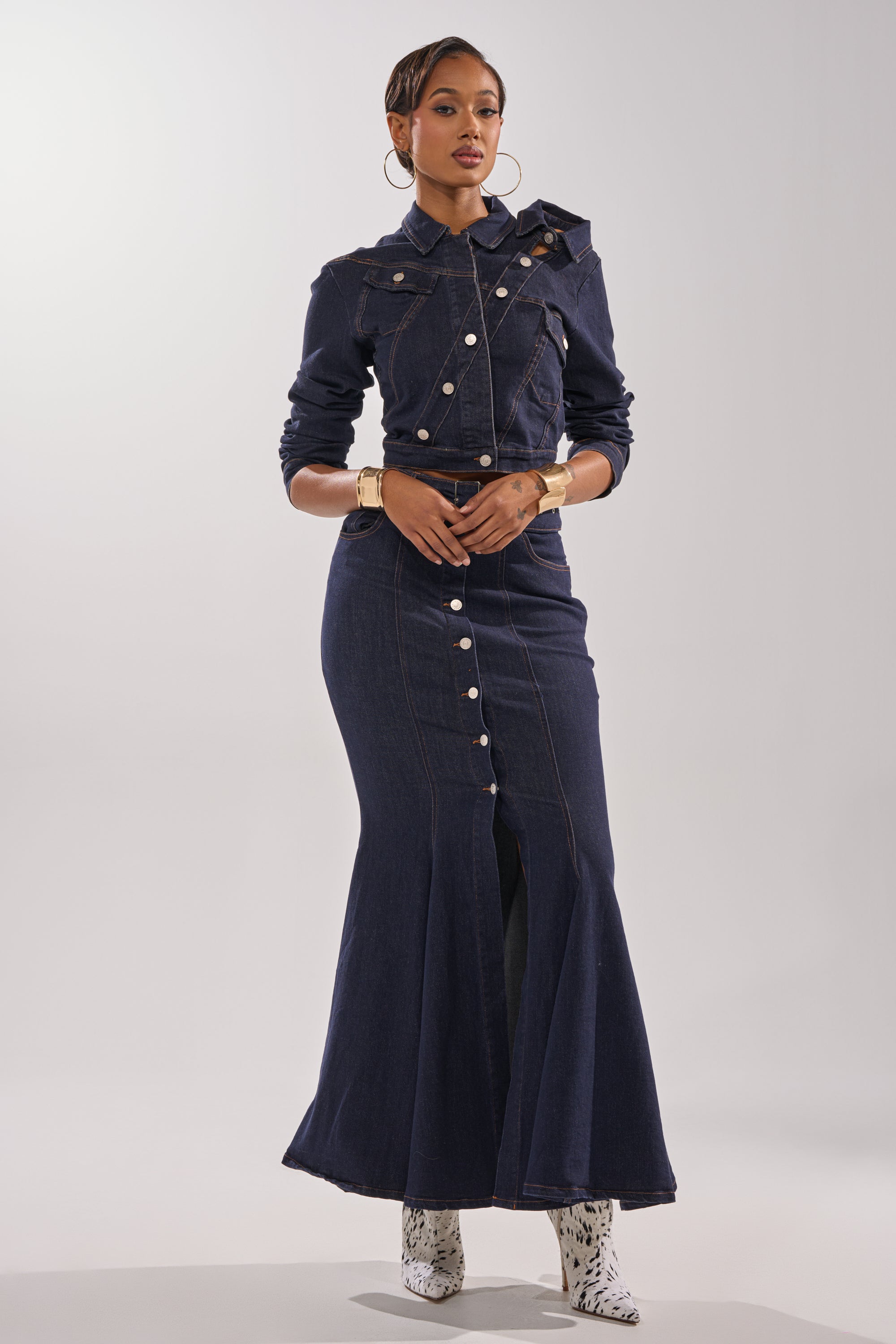 A woman stands against a plain background wearing the YOU GOT ME SPINNING DENIM BUTTON DOWN in indigo wash, styled with long sleeves, white patterned ankle boots, large hoop earrings, and gold bracelets.