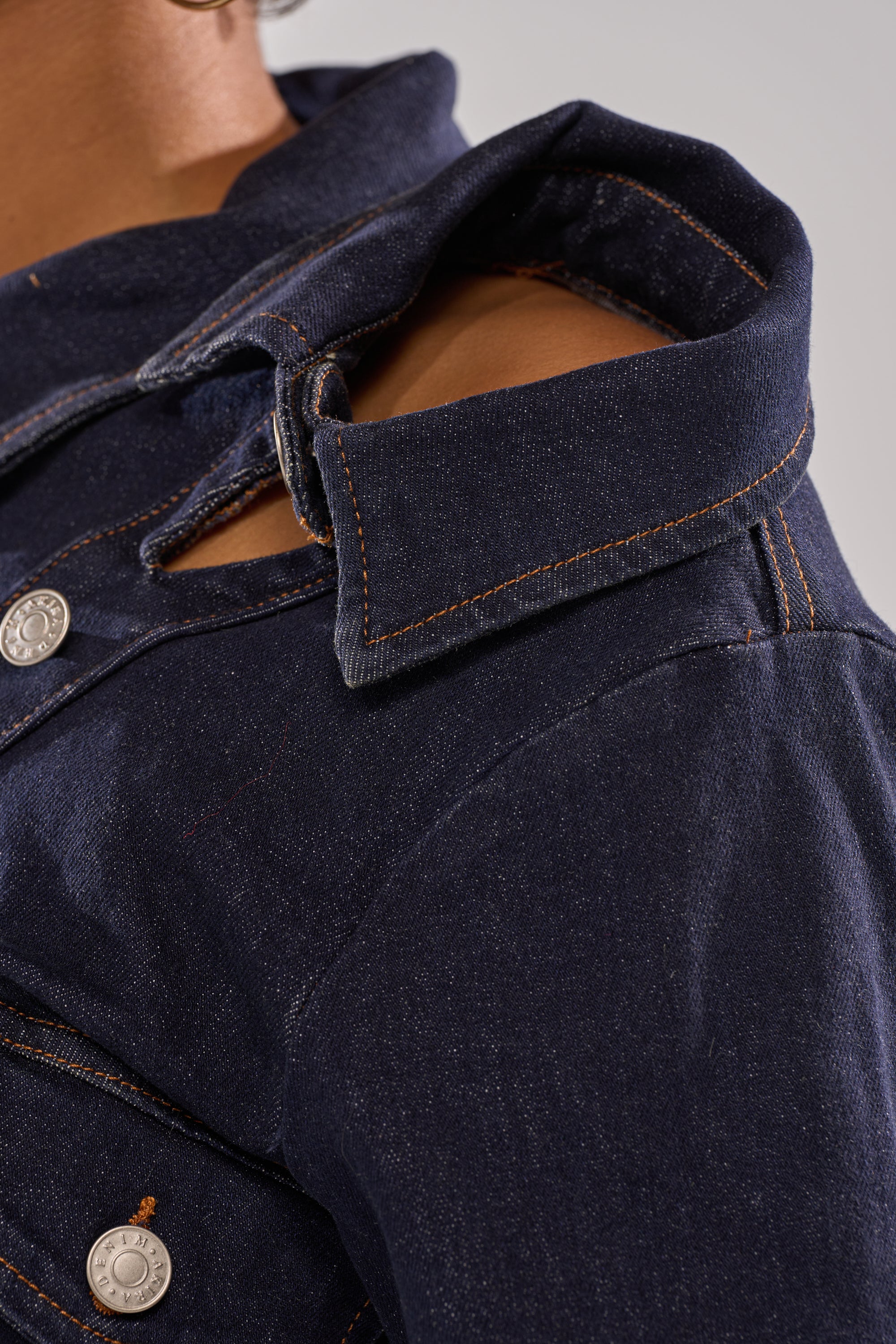 Close-up of a person wearing the YOU GOT ME SPINNING DENIM BUTTON DOWN, featuring orange stitching, an asymmetrical snap-button placket, turned-up collar, and a detached shoulder seam that reveals their bare shoulder.