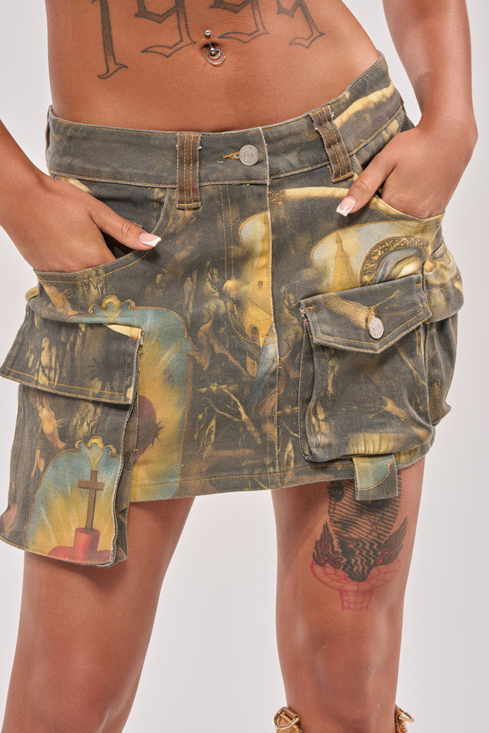 Wearing the AMEN TO THAT MINI SKIRT—a low-rise streetwear piece with multiple pockets and a colorful abstract pattern—this person has visible tattoos on their torso and thigh, hands tucked in the skirt’s pockets. 1