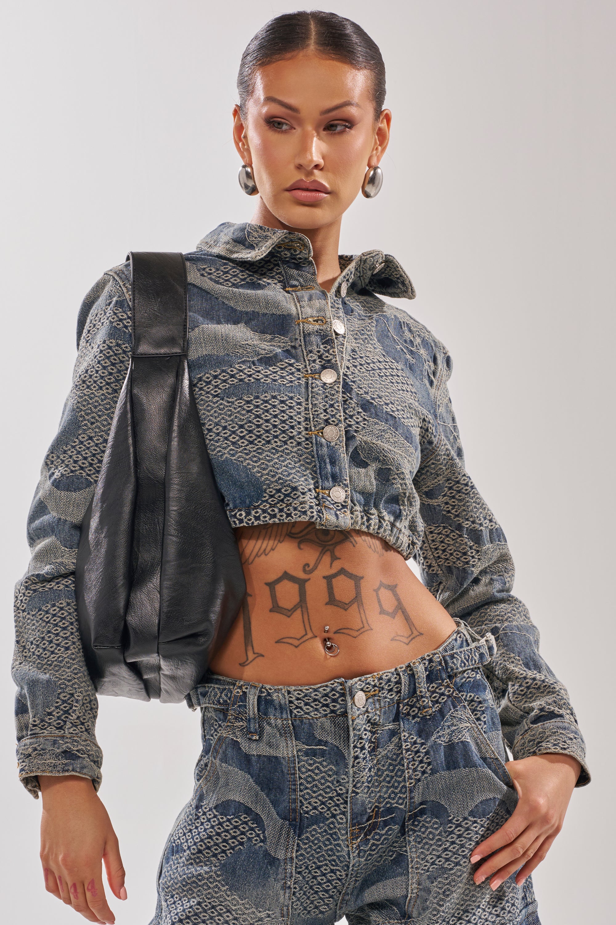 A person with slicked-back hair wears the I THINK YOU WANNA KNOW DENIM BUTTON DOWN and matching jeans, showing a "1998" stomach tattoo. Their look is finished with large hoop earrings and a black shoulder bag.