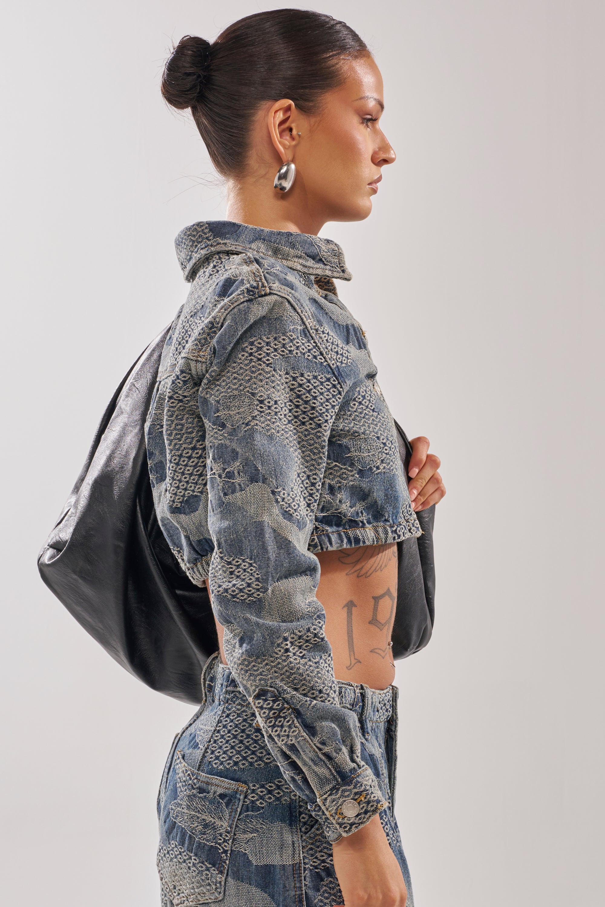 A woman with slicked-back hair and hoop earrings rocks a streetwear look in the I THINK YOU WANNA KNOW DENIM BUTTON DOWN and matching pants. She carries a black bag, revealing a "19" tattoo on her side.