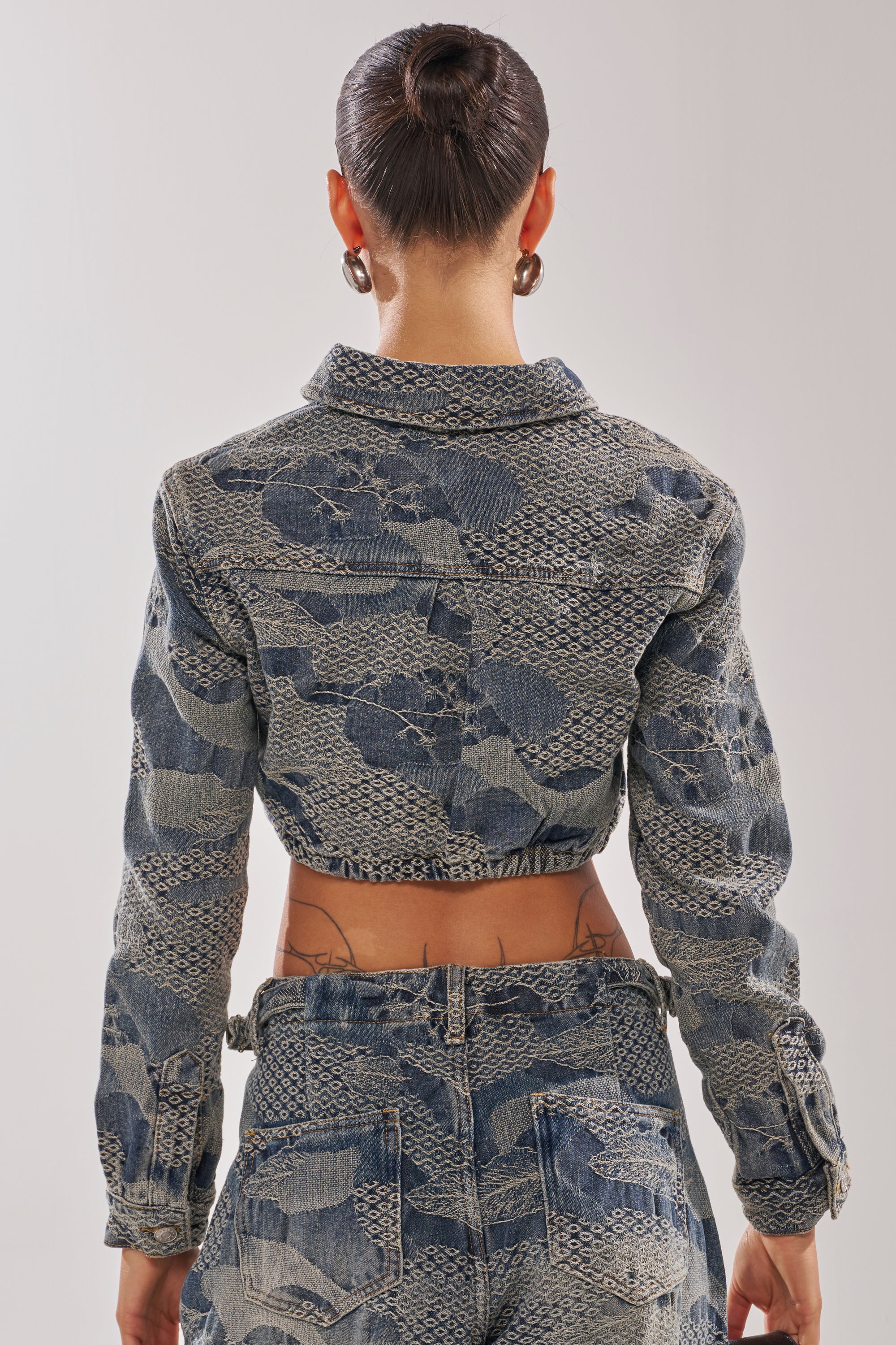 A woman in streetwear, hair in a bun, wears the I THINK YOU WANNA KNOW DENIM BUTTON DOWN with patchwork lace and matching high-waisted jeans. Facing away, she shows tattooed lower back and large hoop earrings.