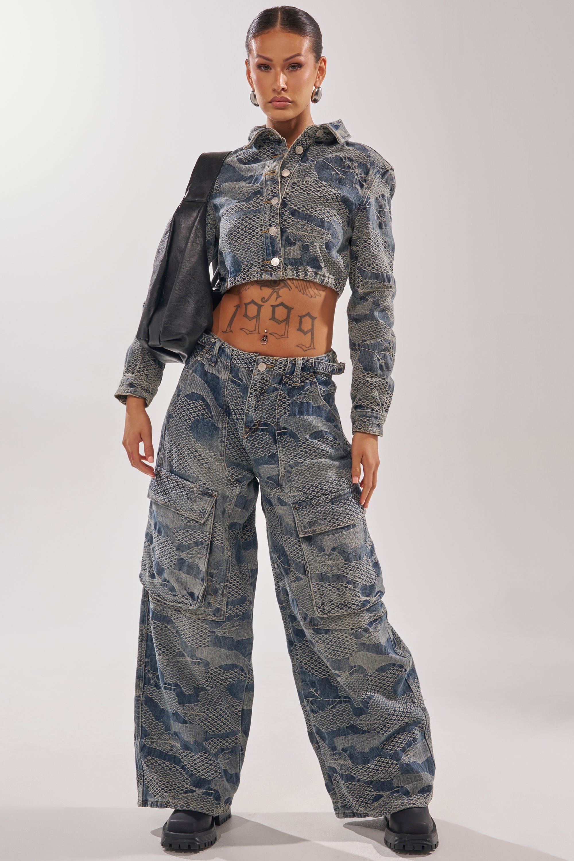 A woman poses confidently in the I THINK YOU WANNA KNOW DENIM BUTTON DOWN, pairing it with wide-leg pants and a black bag, her “1999” tattoo visible on her stomach as she slings a streetwear jacket over one arm.