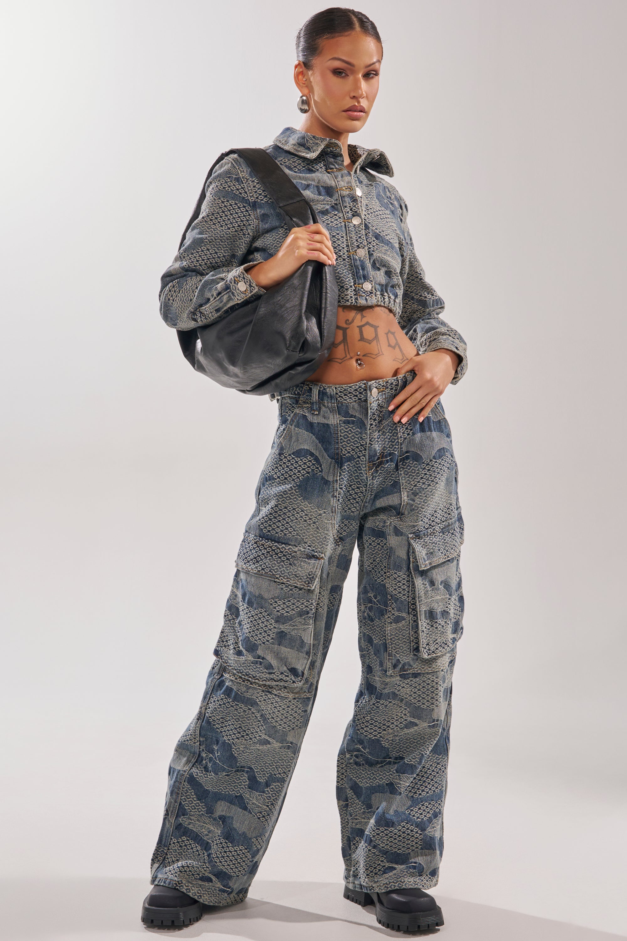 A woman poses confidently in the I THINK YOU WANNA KNOW DENIM BUTTON DOWN and cargo pants, showing her midriff tattoos. She accessorizes with a black shoulder bag and chunky shoes for an edgy, streetwear look.