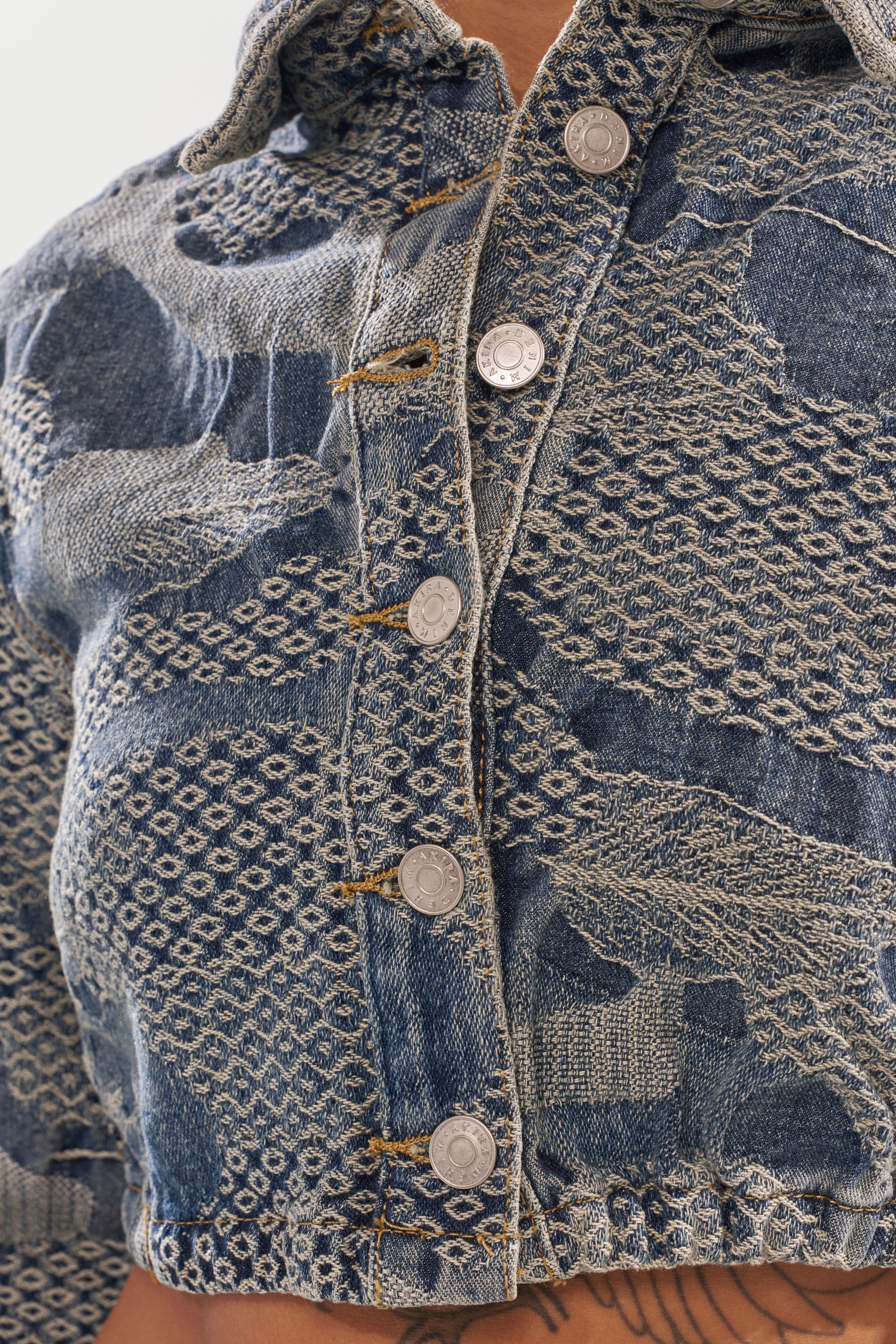 Close-up of the I THINK YOU WANNA KNOW DENIM BUTTON DOWN—a blue cropped jacket featuring textured patterns, silver buttons, frayed stitching, and patchwork lace details for a streetwear edge. Bare skin is visible below the hem.