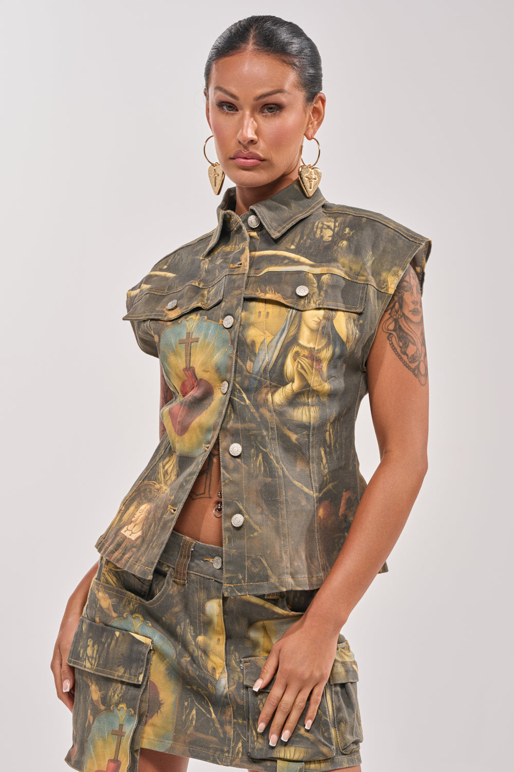 A woman exudes confidence in the AMEN TO THAT BUTTON DOWN denim top with a baroque print and matching skirt, styled with slicked-back hair, bold leaf earrings, and visible arm tattoos for a striking streetwear festival look. 0