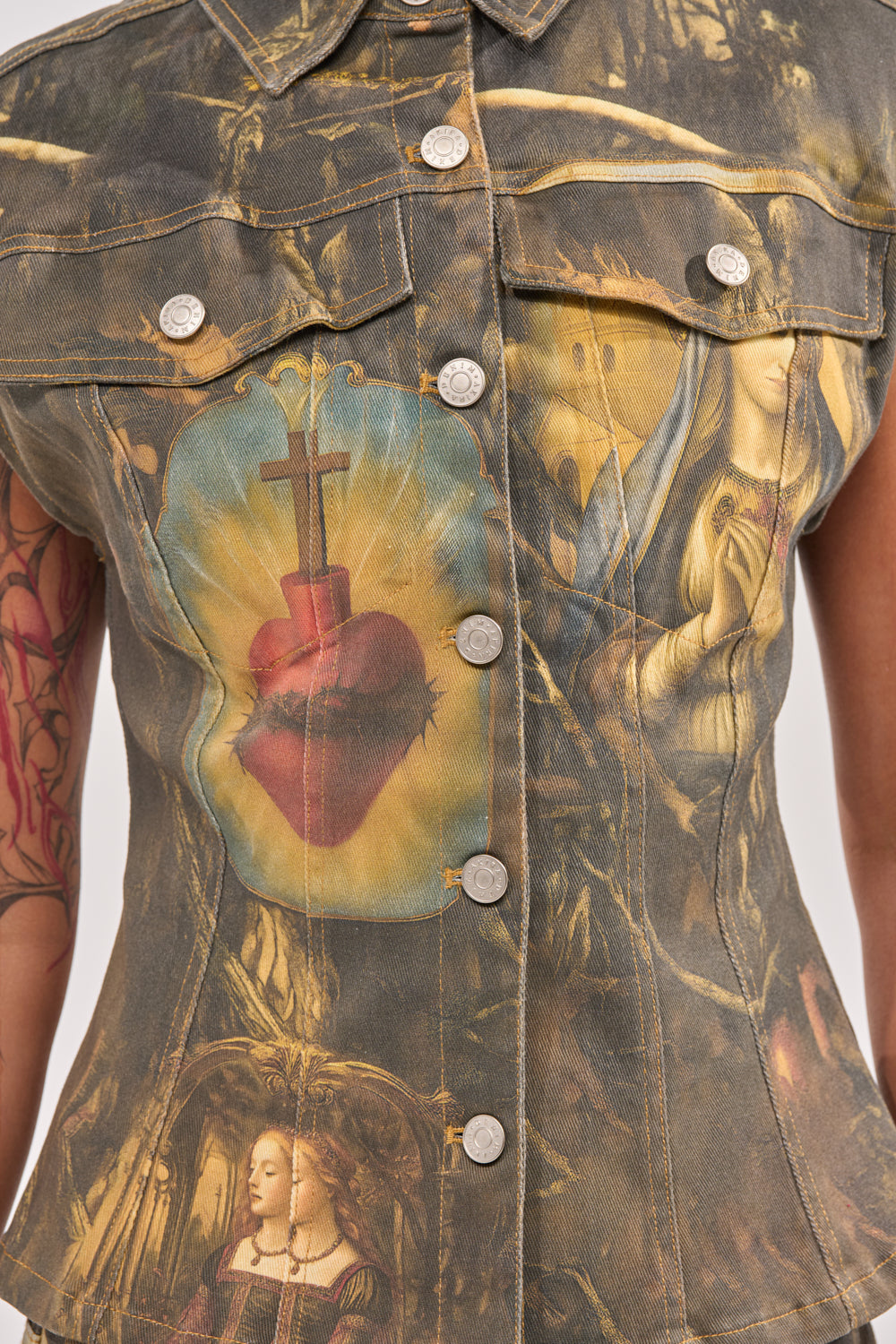 The AMEN TO THAT BUTTON DOWN is a sleeveless denim top with baroque, religious-inspired prints—like sacred hearts and Renaissance women—and silver front buttons for an edgy, streetwear-ready festival look. 10