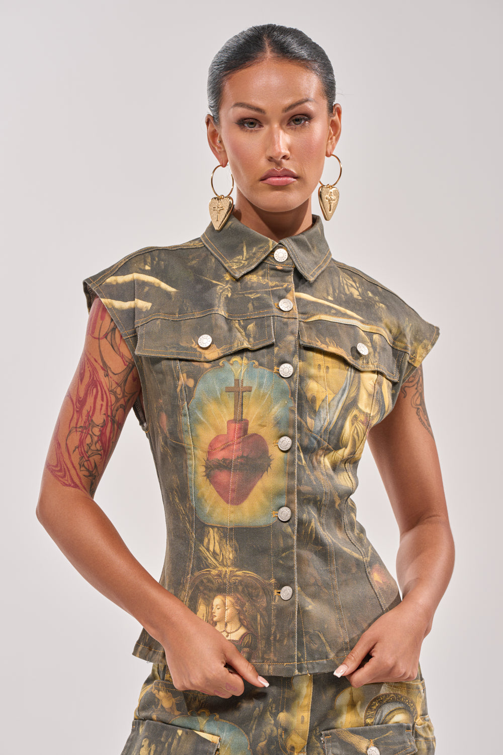 A woman with slicked-back hair wears the AMEN TO THAT BUTTON DOWN, a sleeveless vest with baroque religious art and a flaming heart motif, styled with large heart-shaped earrings against a plain background. 2