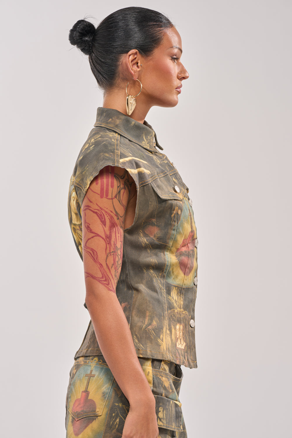 A woman with dark hair in a bun and gold earrings stands in profile, wearing the AMEN TO THAT BUTTON DOWN denim top and matching shorts—an eye-catching festival outfit. She has a large red tattoo on her upper arm. 4