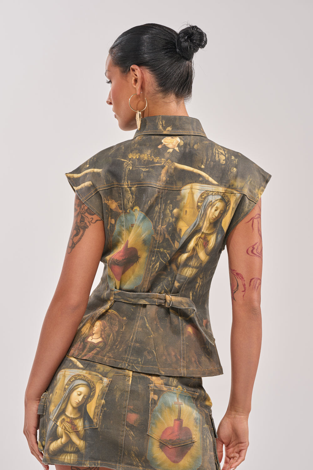 A woman with dark hair in a bun models the AMEN TO THAT BUTTON DOWN—a sleeveless, belted jacket and matching skirt set with religious artwork and heart motifs—posing turned away to display the back and side of her festival outfit. 5