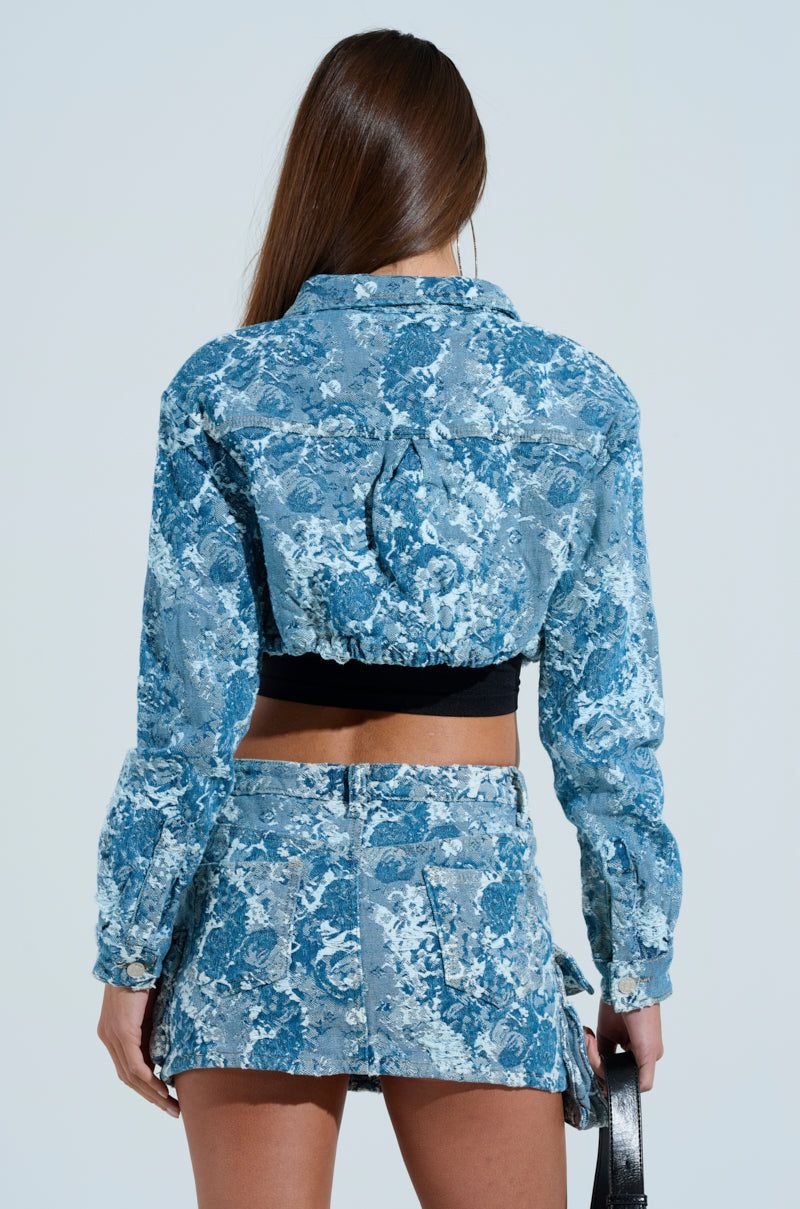 A person with long hair is seen from behind, wearing the DAY N NIGHT CROPPED DENIM BUTTON DOWN and a matching mini skirt. The outfit has a blue and white pattern with a denim-like look enhanced by distressed stitching, and the individual holds a small black handbag.