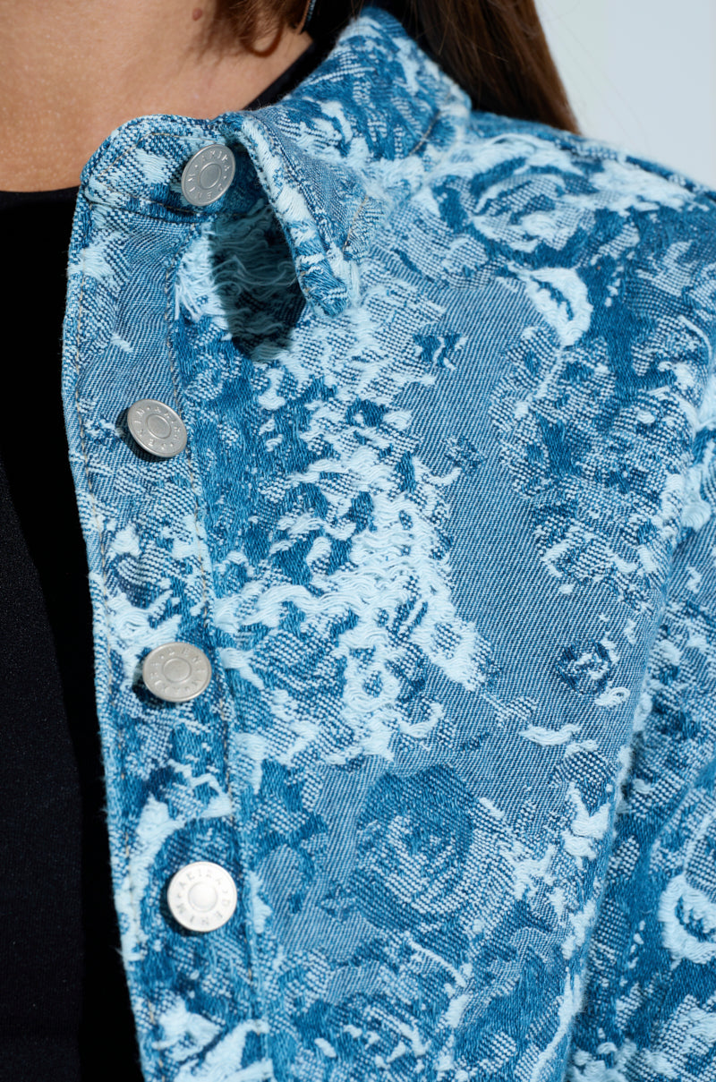 A close-up shows a person wearing the DAY N NIGHT CROPPED DENIM BUTTON DOWN over a black top, featuring silver buttons, distressed stitching, and a blue and white floral design that accentuates the fabric's texture.