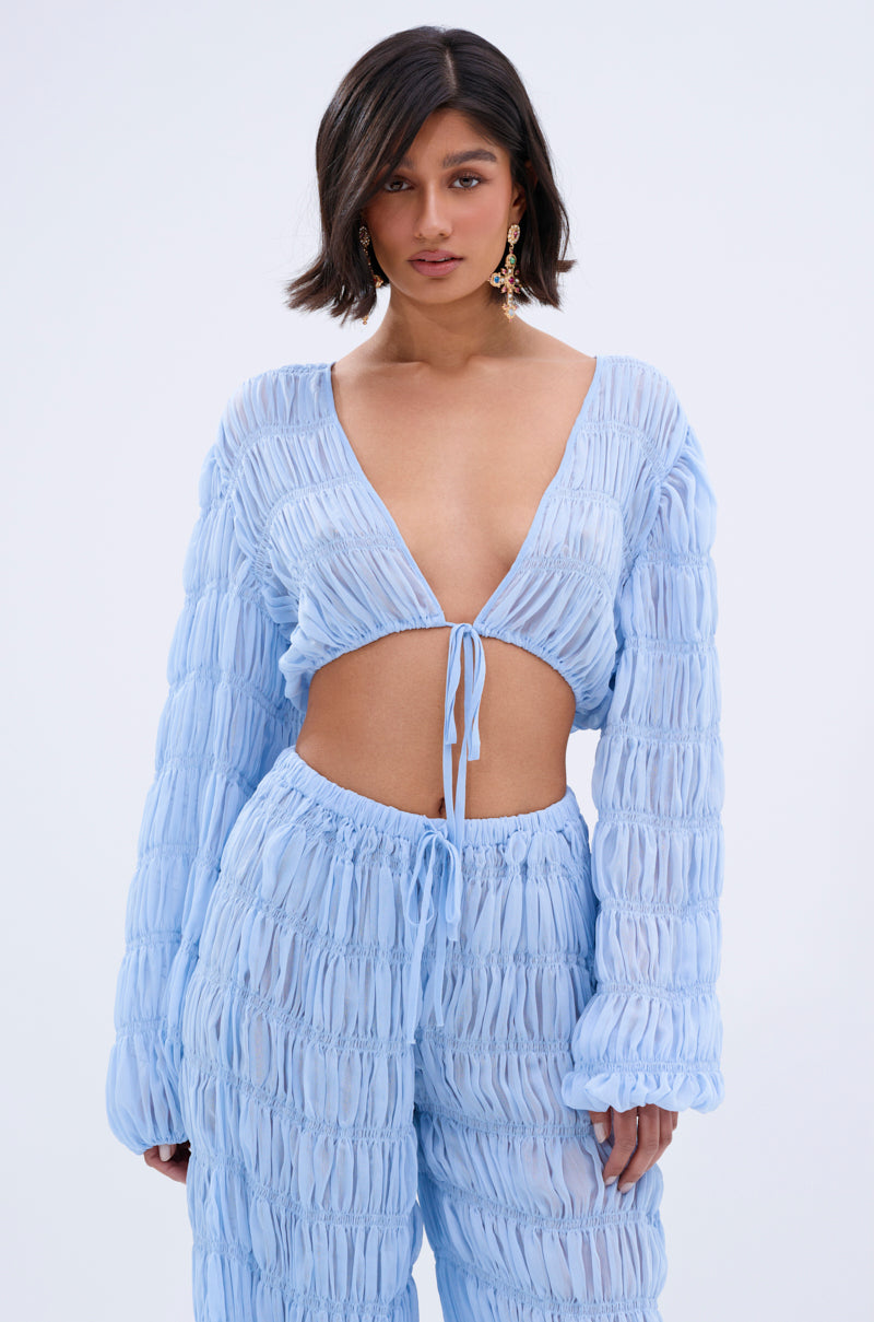 A woman with short dark hair wears the ICED OUT TIE FRONT TOP, a light blue, textured piece with a deep V neckline and boho vibe. She pairs it with matching high-waisted pants and stands confidently against a plain background.