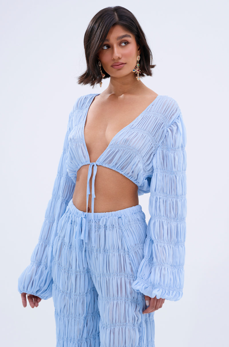 Wearing the ICED OUT TIE FRONT TOP, a light blue, textured, long-sleeve crop with matching high-waisted pants, a woman with shoulder-length dark hair and dangling earrings poses against a plain background.