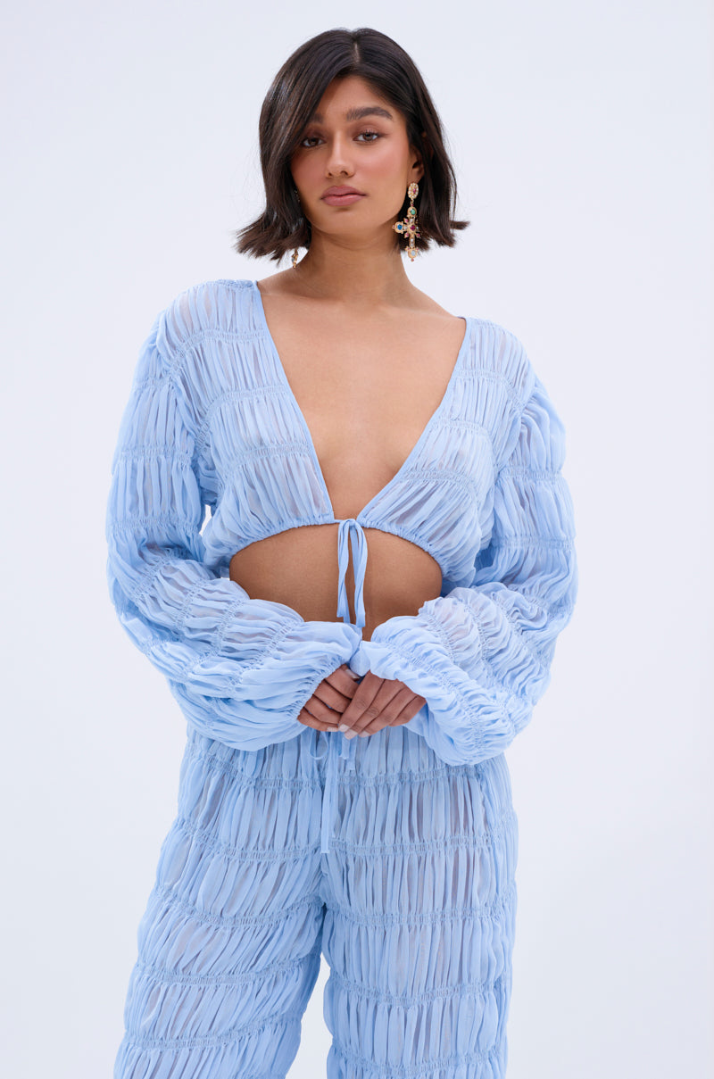 Wearing the ICED OUT TIE FRONT TOP, a person with short dark hair and dangling earrings stands against a plain background. The light blue, sheer, textured two-piece features long sleeves and a deep V-neck with a tie-front design.