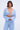 Wearing the ICED OUT TIE FRONT TOP, a person with short dark hair and dangling earrings stands against a plain background. The light blue, sheer, textured two-piece features long sleeves and a deep V-neck with a tie-front design.