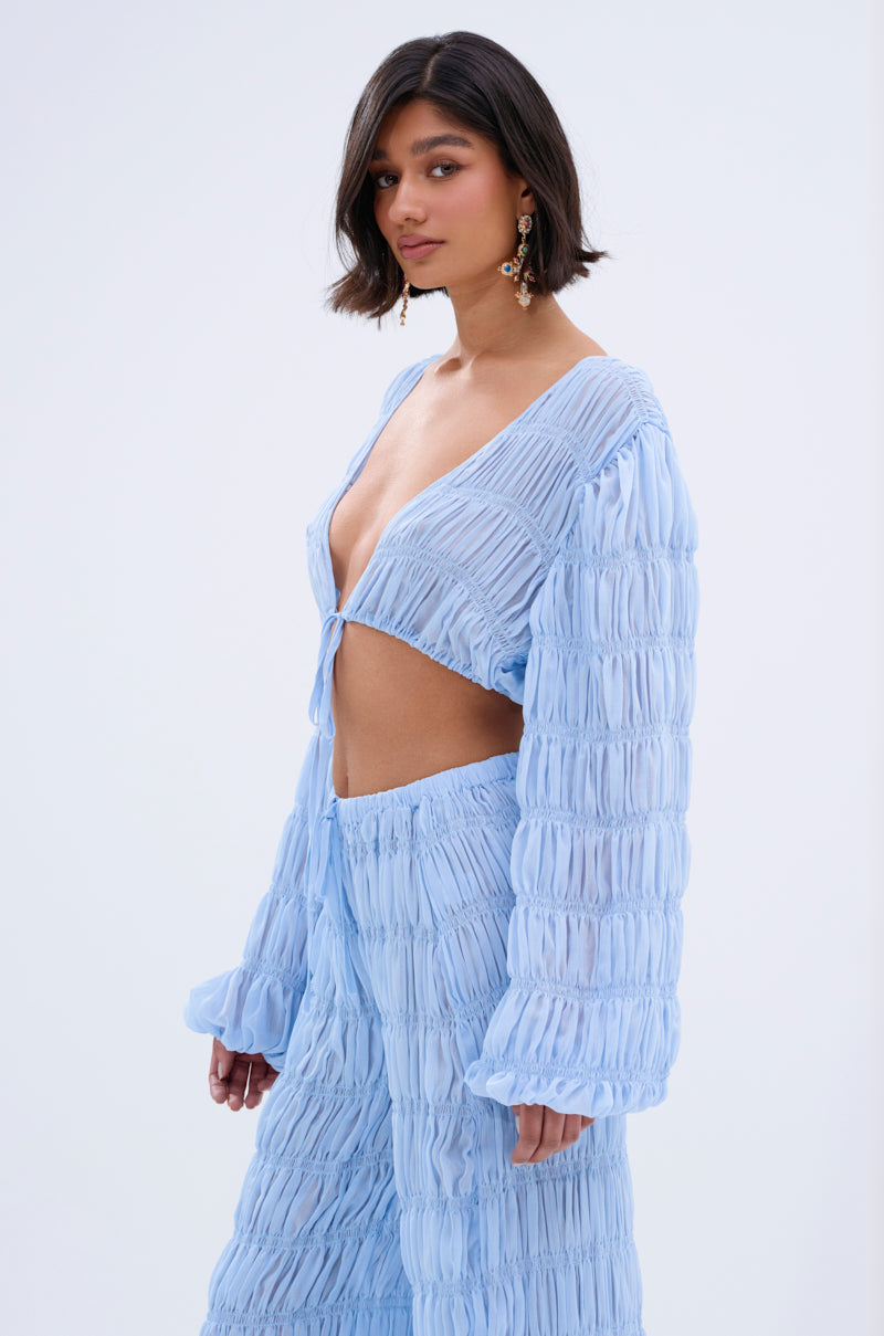 A woman with short dark hair models the ICED OUT TIE FRONT TOP, a light blue, textured blouse with wide sleeves and a deep V-neck, styled in a boho vibe. She stands against a plain light background, facing the camera with a neutral expression.
