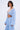 A woman with short dark hair models the ICED OUT TIE FRONT TOP, a light blue, textured blouse with wide sleeves and a deep V-neck, styled in a boho vibe. She stands against a plain light background, facing the camera with a neutral expression.