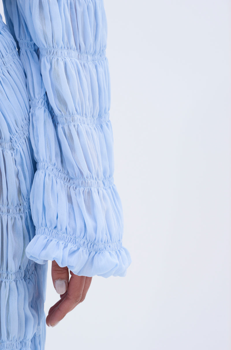 A close-up of a person's arm wearing the ICED OUT TIE FRONT TOP in light blue sheer chiffon with ruched texture. The person has light-colored nail polish, set against a plain, light background.