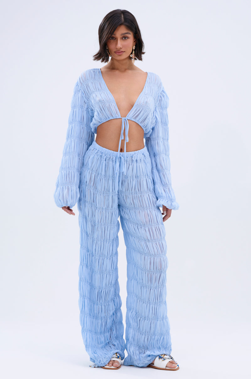 A woman with short dark hair stands against a white background wearing the ICED OUT PALAZZO PANT—light blue, textured, loose chiffon pants styled with a matching deep V-neck tie-front top and sandals for a chic boho vibe.
