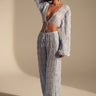 A woman models the ICED OUT PALAZZO PANT, a light blue textured wide-leg pant, paired with a matching long-sleeve cropped top featuring a deep V-neck and tie front. She completes the look with sandals and statement earrings against a neutral backdrop.