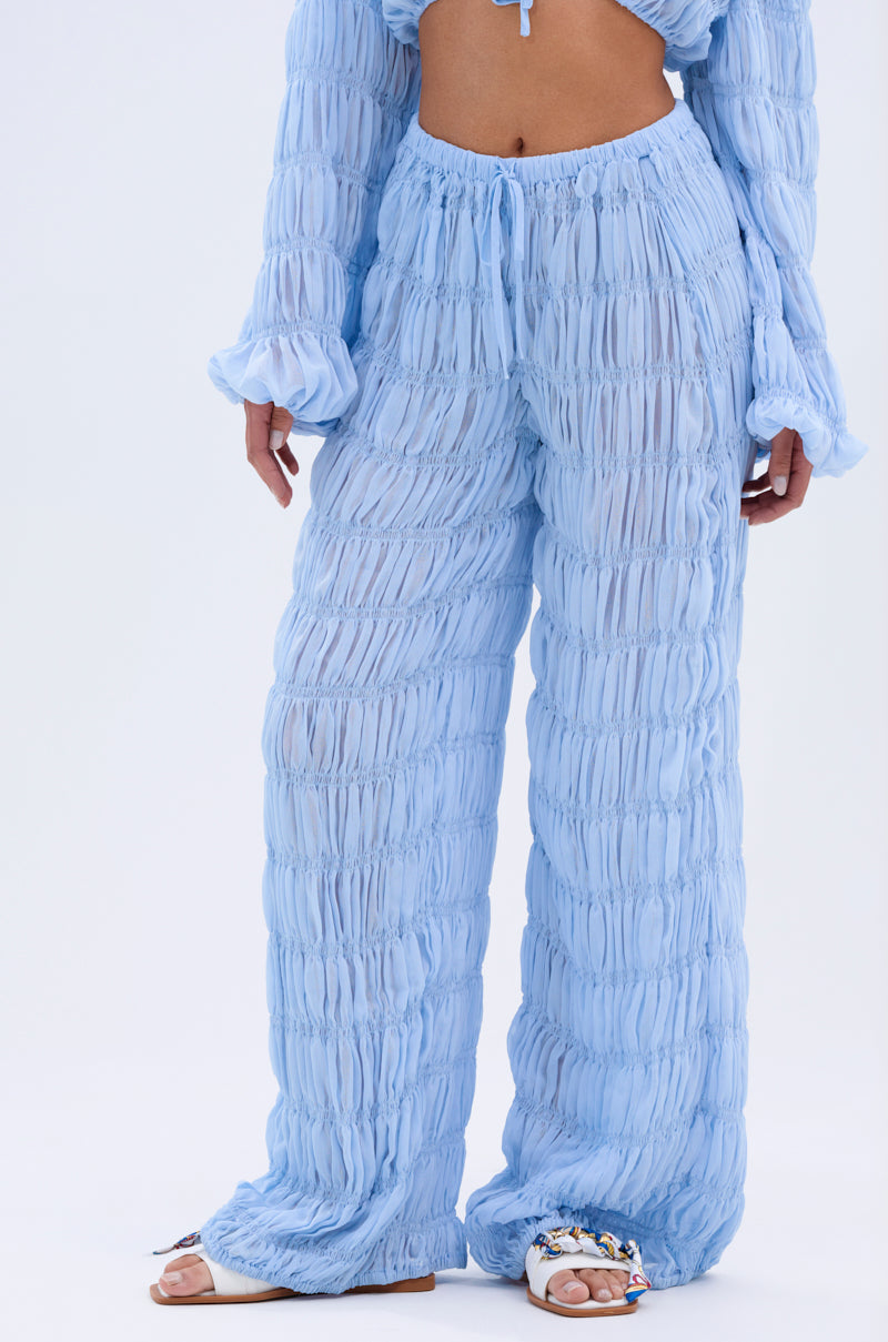A person models the ICED OUT PALAZZO PANT in light blue chiffon with a matching top, revealing some midriff. The sheer pants have an elastic waistband and are styled with flat, decorative open-toe sandals.