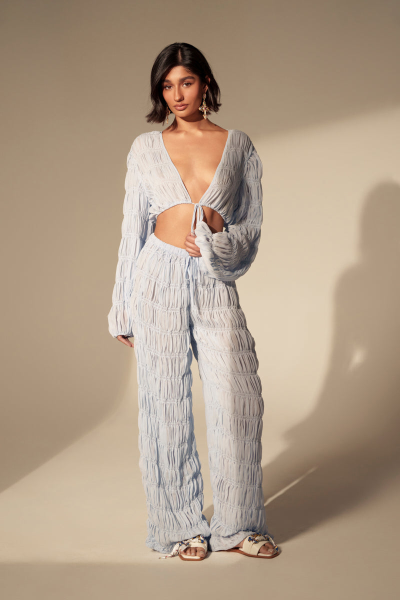 A woman with short dark hair poses against a neutral background in the ICED OUT PALAZZO PANT and a light blue textured deep V-neck top, styled with sandals and statement earrings for a chic, modern look.