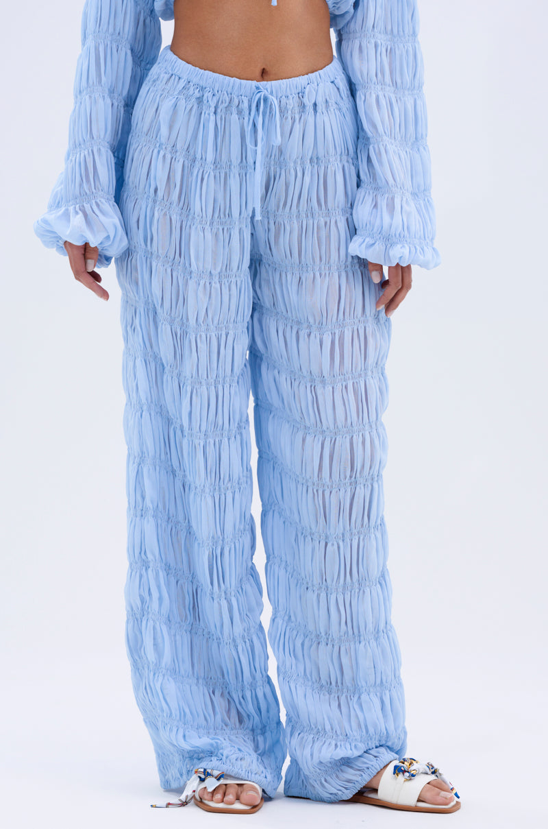 A person models the ICED OUT PALAZZO PANT in light blue sheer textured chiffon with a drawstring waist, paired with a matching long-sleeve top and embellished white slide sandals, all set against a plain white background.