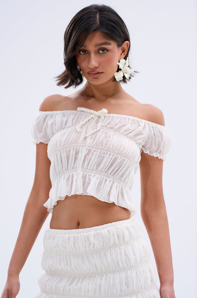 A woman with short dark hair models the FLIRT WITH ME OFF THE SHOULDER BLOUSE, which features a ruffled hem, paired with a matching skirt and large white flower earrings against a plain light background.