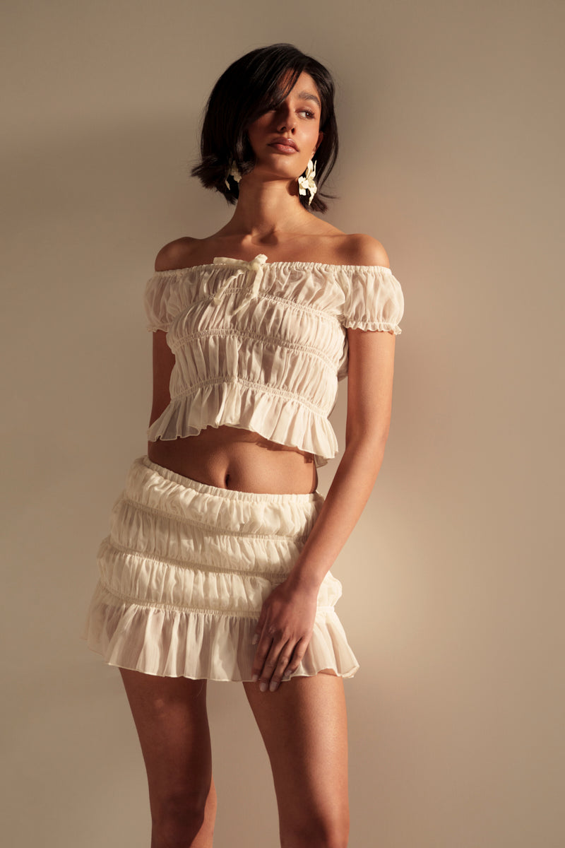 Wearing the FLIRT WITH ME OFF THE SHOULDER BLOUSE in sheer white crepe and a matching ruffled hem mini skirt, a person poses against a neutral background with one hand on their thigh and the other arm relaxed.