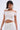 A woman with short dark hair models the FLIRT WITH ME OFF THE SHOULDER BLOUSE, which features a ruffled hem, paired with a matching skirt and large white flower earrings against a plain light background.