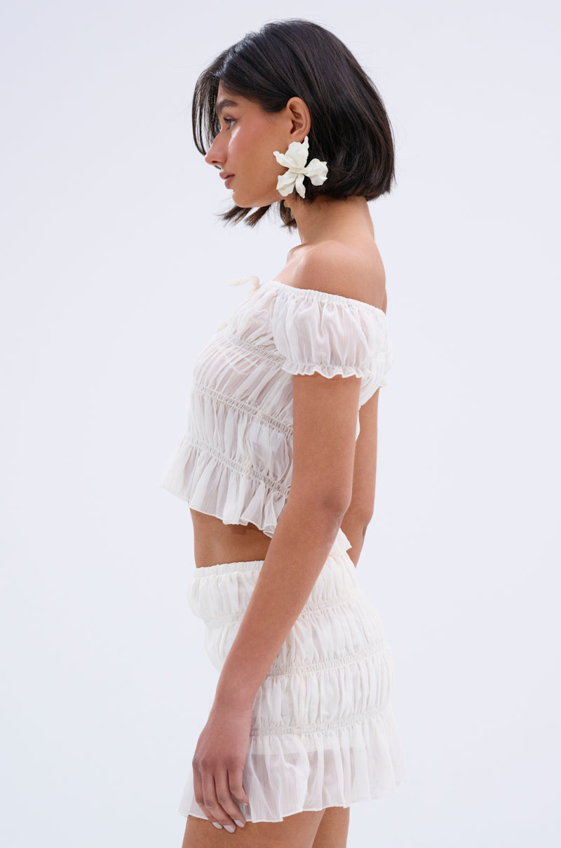 A woman with short dark hair stands in profile, wearing the FLIRT WITH ME OFF THE SHOULDER BLOUSE and a matching ruffled skirt. She accessorizes with a white flower earring against a plain light background.